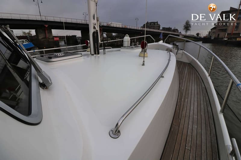 De foto toont het voordek van een boot met een wit dek, houten zijdekken en een reling, met een brug op de achtergrond.