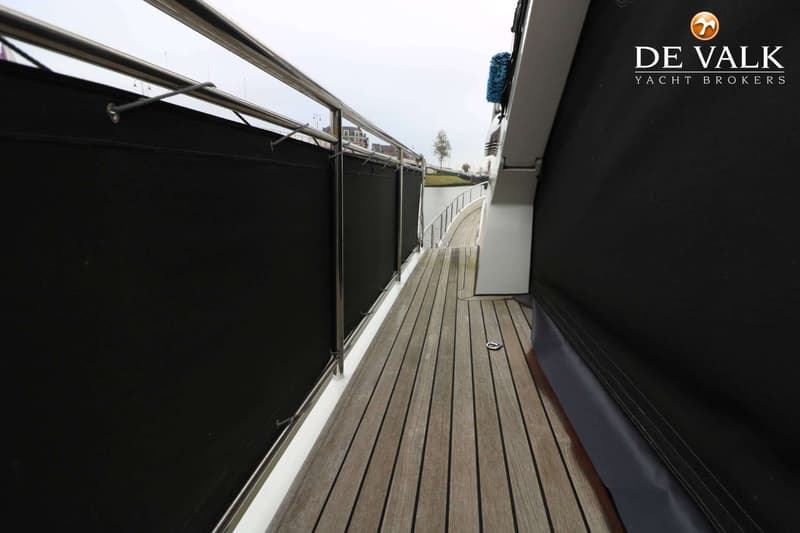 Een houten dek met zwarte zeilen aan de zijkanten en een reling, met een waterweg en gebouwen op de achtergrond.