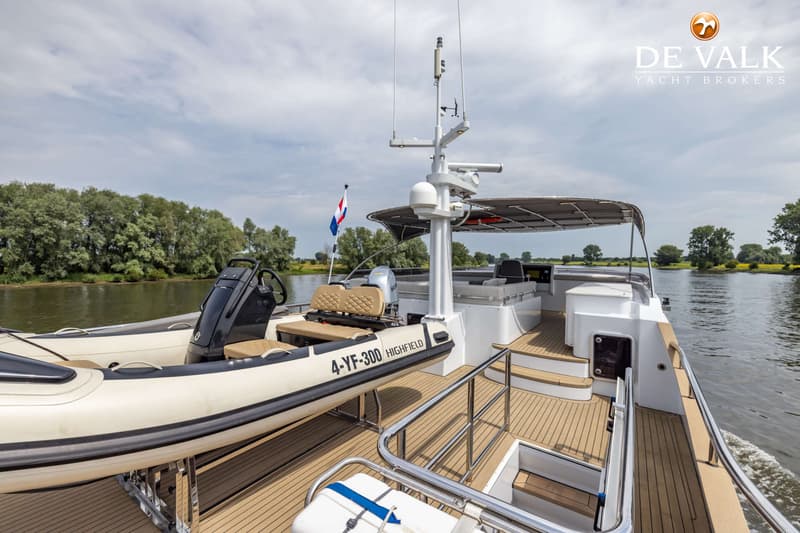 Een witte motorboot met een bijboot aan dek vaart op een rivier met bomen langs de oevers onder een bewolkte lucht.