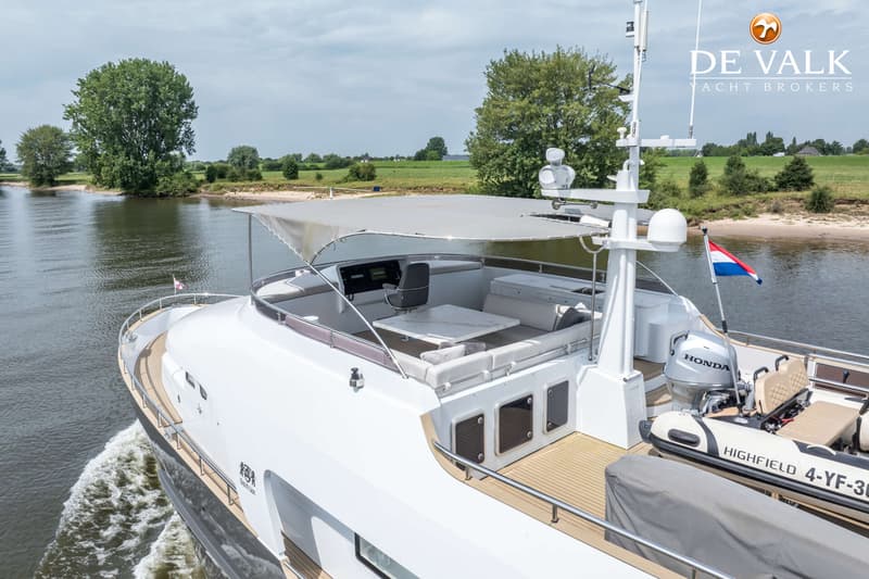Een witte motorboot vaart op een rivier met groene oevers, met een bijboot en een Nederlandse vlag achterop.