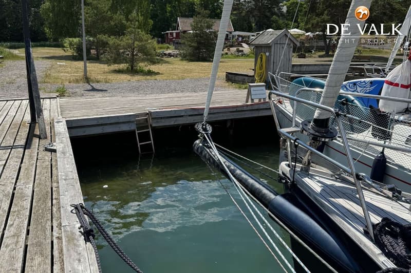 De foto toont het teakhouten dek van een zeilboot met twee stuurwielen, lieren en een buitenboordmotor aan de spiegel.