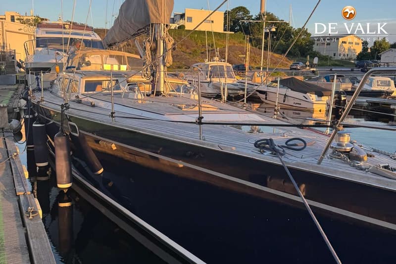Een bovenaanzicht van het teakhouten dek van een zeilboot met luiken, lieren en een mast, naast een andere boot.