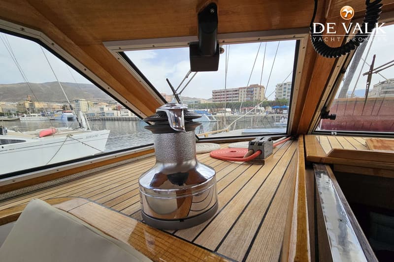Een interieurzicht van een boot met houten afwerking, een lier, en uitzicht op een haven met andere boten en gebouwen.