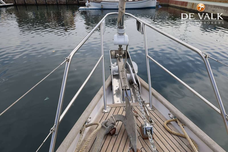 De foto toont de boeg van een zeilboot met een anker, ankerketting, touwen en een reling, liggend in het water.