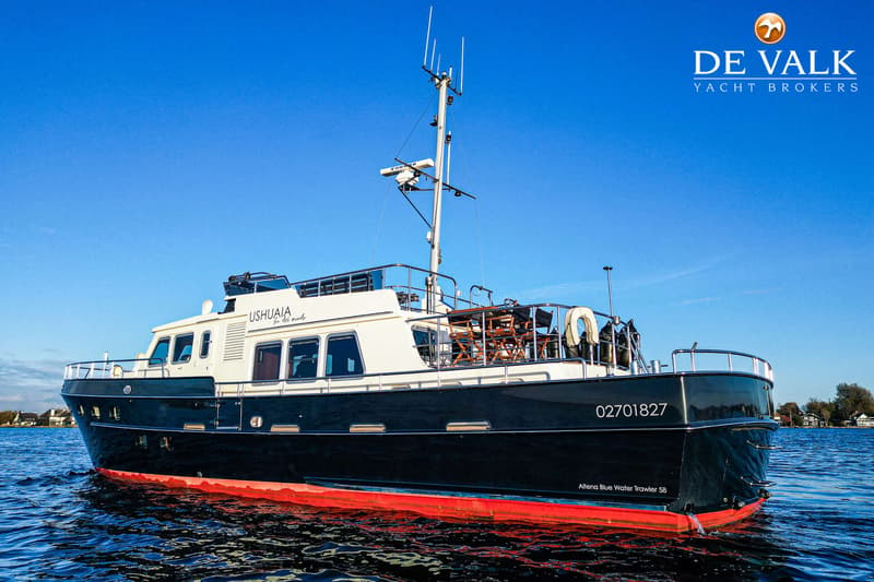 Een donkerblauwe boot met rode onderkant en witte opbouw, genaamd "USHUAIA", vaart op rustig water onder een helderblauwe…