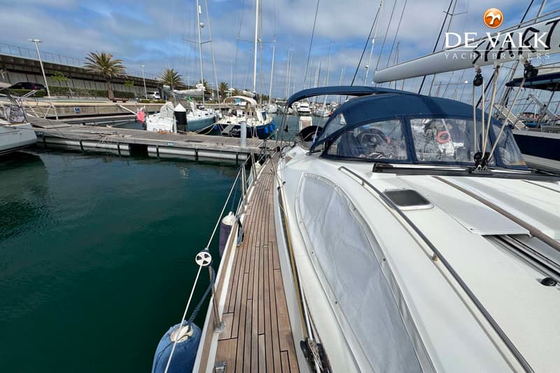 Een witte zeilboot met een blauwe buiskap ligt afgemeerd in een jachthaven, met een houten dek en andere boten op de…