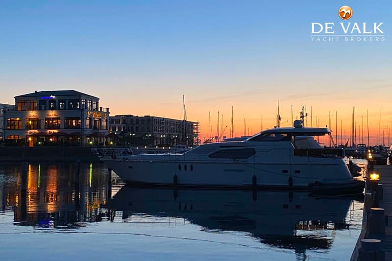 Een motorjacht ligt aangemeerd in een haven bij zonsondergang, met gebouwen en andere boten op de achtergrond.