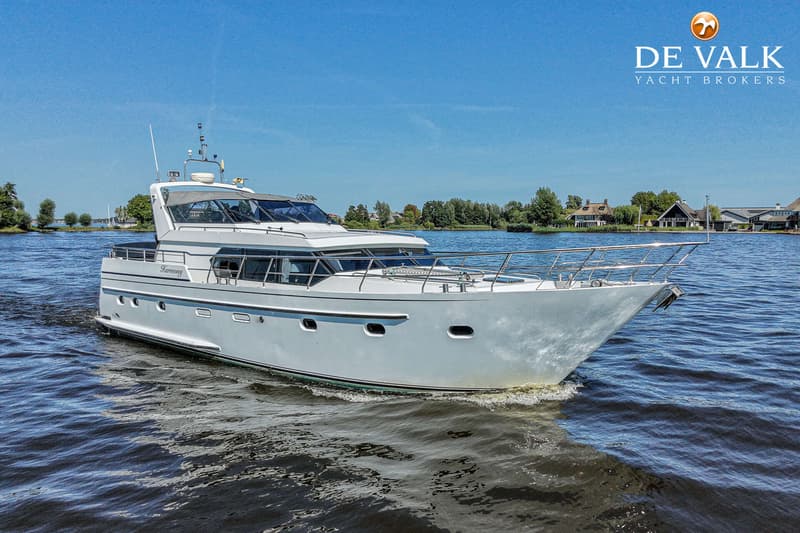 Een wit motorjacht met de naam "Harmony" vaart op een waterweg met bebouwing en bomen op de achtergrond onder een blauwe…