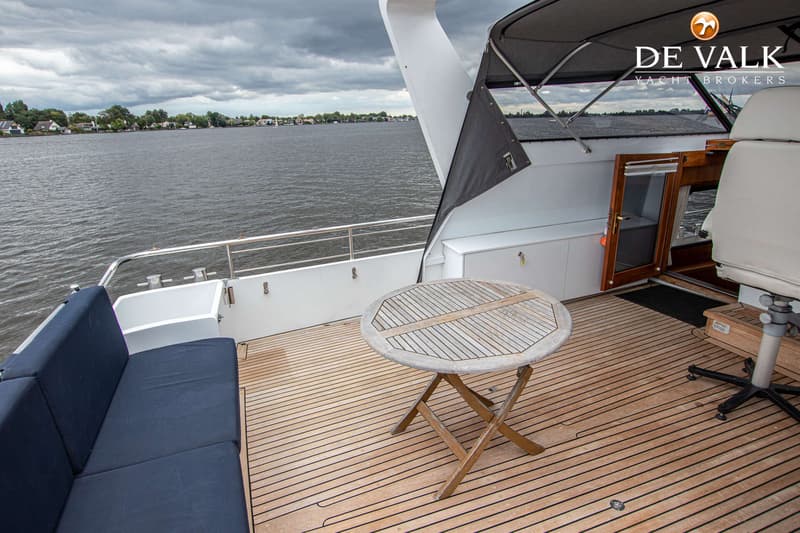 Een teakhouten achterdek met een ronde houten tafel, een blauwe bank en een open deur naar de kajuit, met water op de…