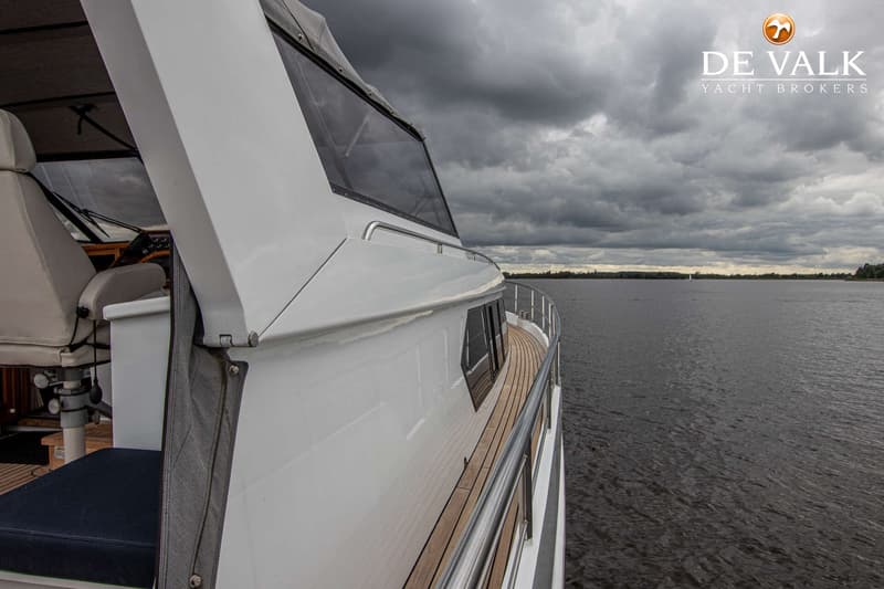 Een witte boot met een houten dek en reling vaart op water onder een bewolkte lucht, met land in de verte.
