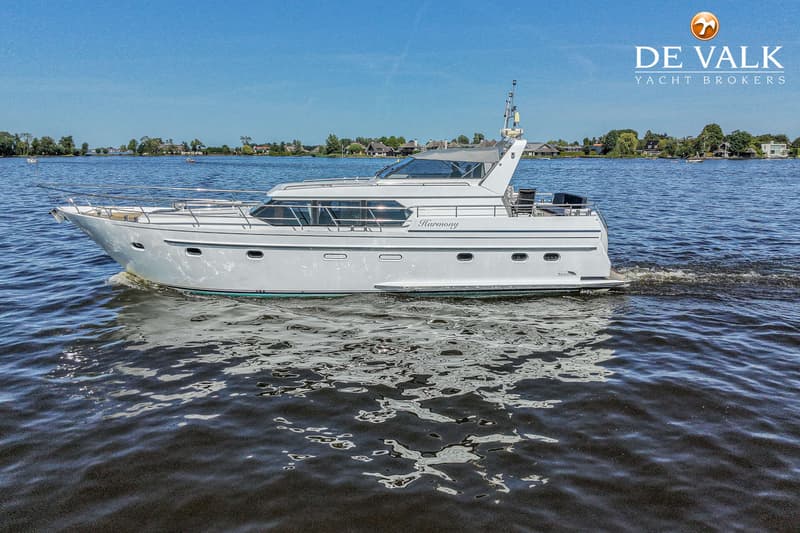 Een witte motorjacht met de naam "Harmony" vaart op een waterweg met beboste oevers onder een blauwe lucht.