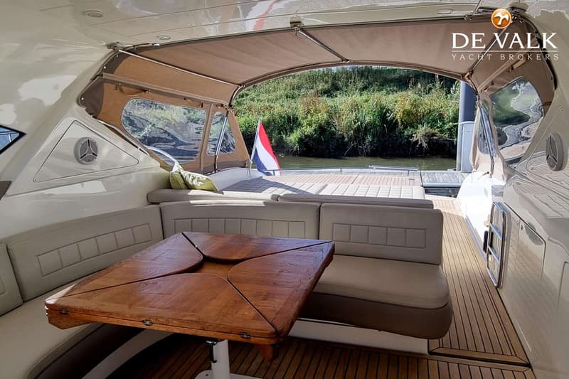 De foto toont het achterdek van een boot met een houten tafel, banken, een Nederlandse vlag en een uitzicht op water en…