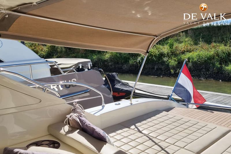 Een deel van een boot met een zonnescherm, kussens, een Nederlandse vlag en een waterkant met groen. — Conam 58 Sport HT