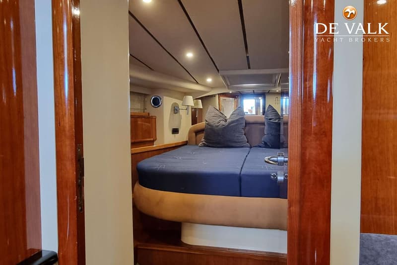 Een slaapkamer in een boot met een donkerblauw bed, kussens, houten wanden en een patrijspoort. — Conam 58 Sport HT