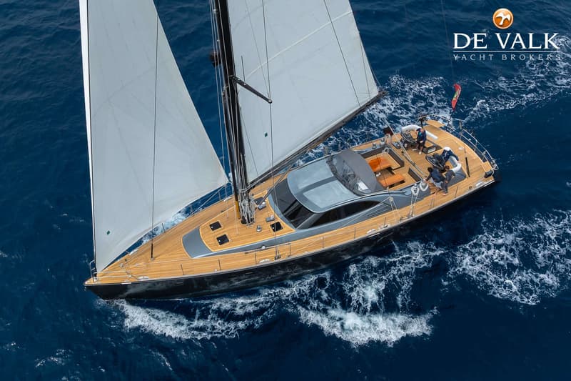 Een zeilboot met twee witte zeilen en een teakhouten dek vaart op open water met vier personen aan boord.