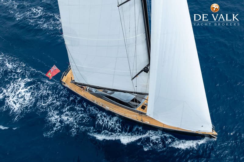 Een zeilboot met twee witte zeilen en een houten dek vaart op blauw water, met een vlag aan de achtersteven.