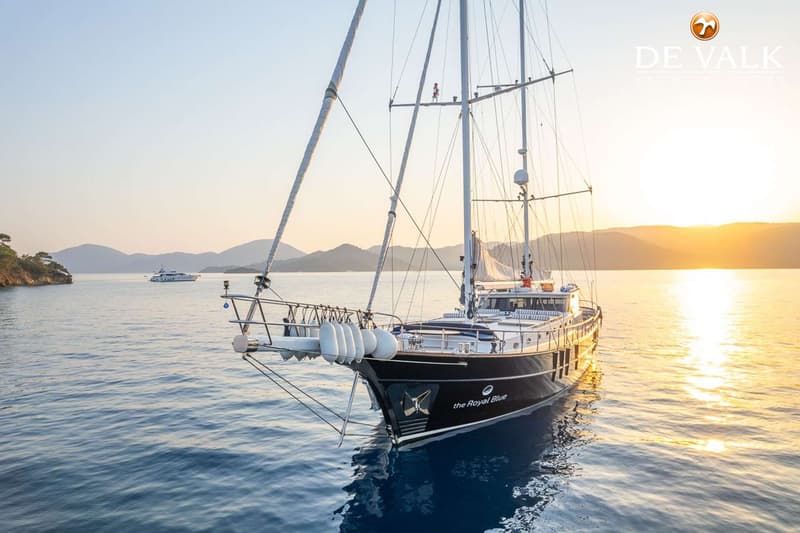 Een donkerblauwe zeilboot met de naam "the Royal Blue" ligt op het water bij zonsondergang, met bergen op de achtergrond.