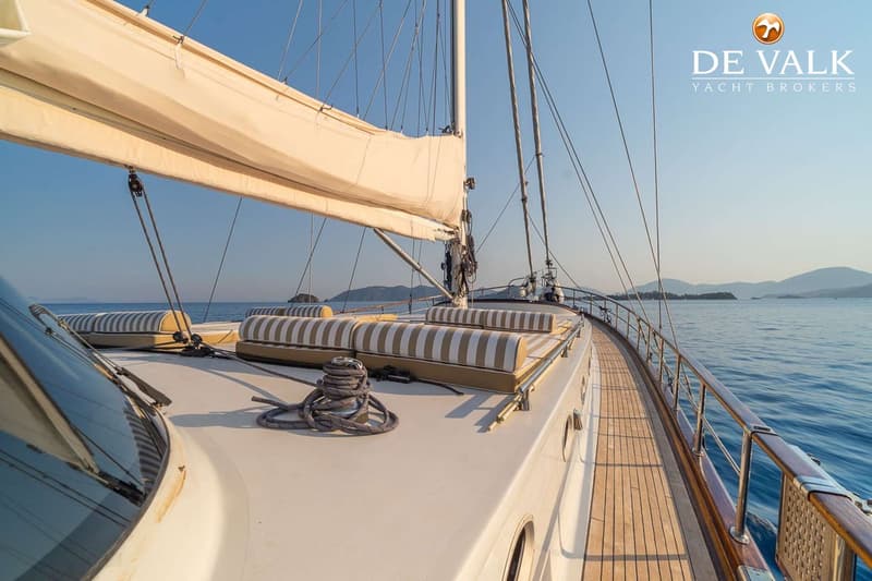 Een zeilboot met een opgerold zeil, ligkussens en een houten dek, varend op zee met bergen op de achtergrond. — Gullet 27