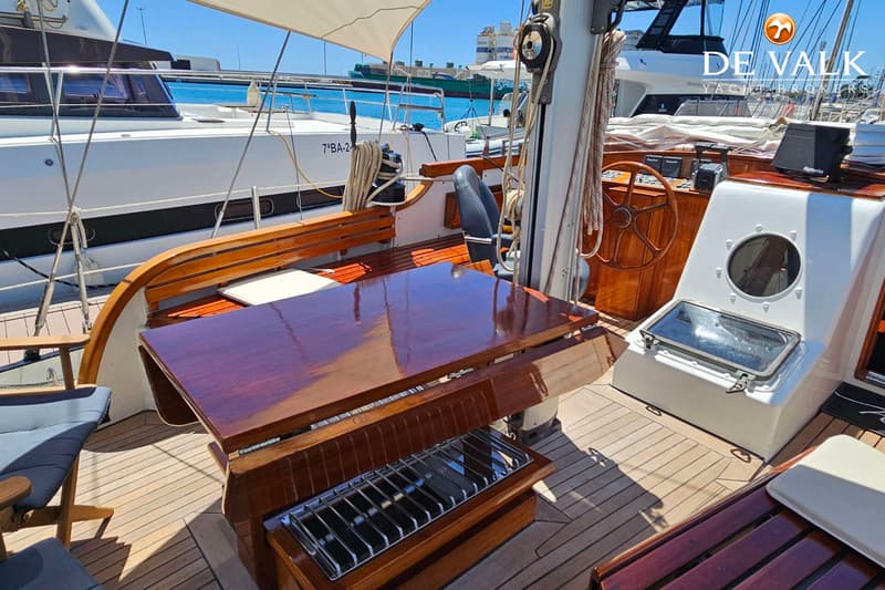 Een houten dek met een glanzende houten tafel, banken en een stuurwiel, met een witte boot op de achtergrond.