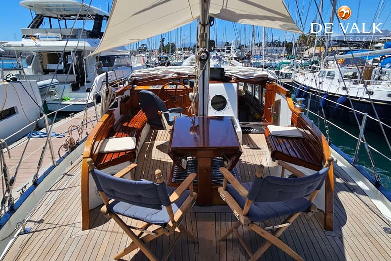 Een houten dek met een tafel en twee stoelen, omringd door houten banken, onder een wit zeil in een jachthaven.