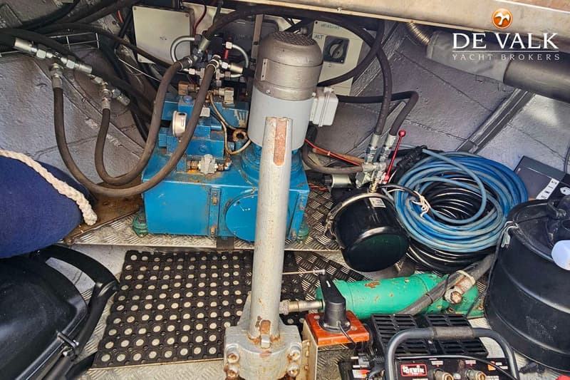 Een detailfoto van een machinekamer met een blauwe hydraulische pomp, slangen, kabels en een rubberen mat op de vloer.