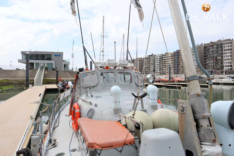 Een grijze boot ligt afgemeerd aan een steiger, met gebouwen en andere boten op de achtergrond. — Waarschip 1076 Custom