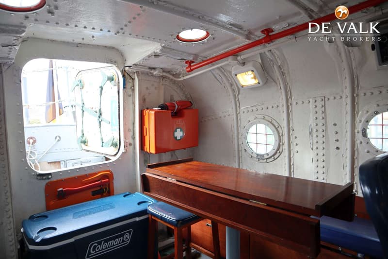 Een interieur van een boot met een houten tafel, een blauwe koelbox, een oranje EHBO-doos en patrijspoorten.