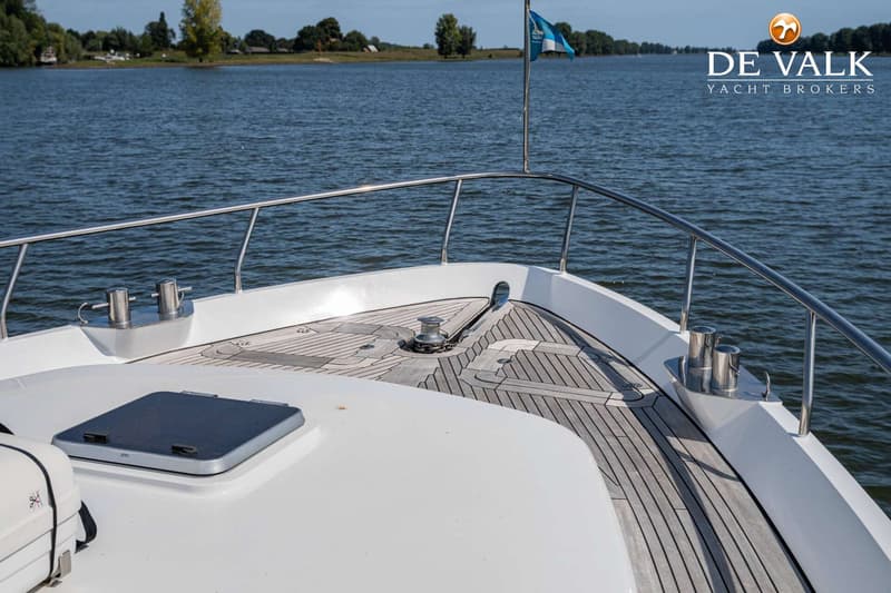 De foto toont het voordek van een boot met teakhouten dek, een ankerlier, reling, bolders en een luik.