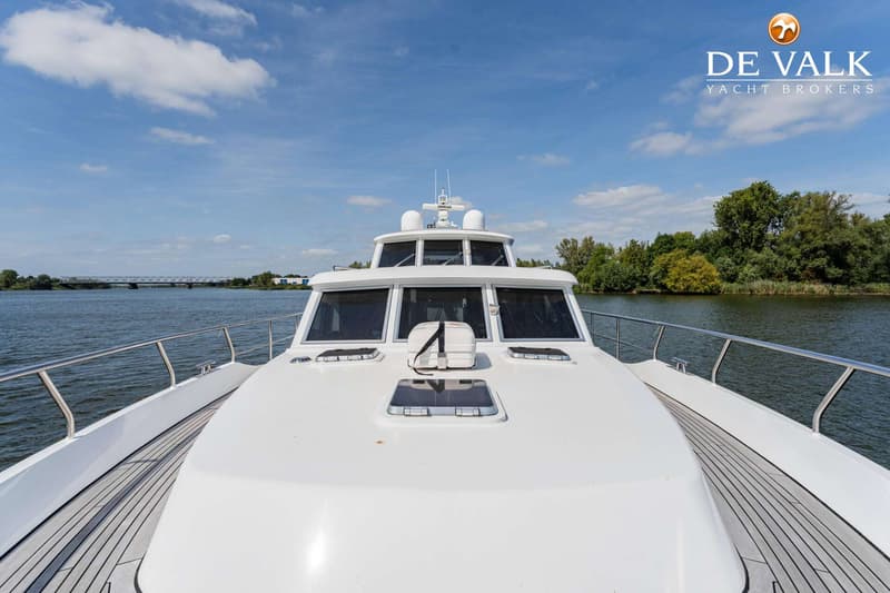 Een witte motorboot vaart op een rivier met een brug en bomen op de achtergrond onder een blauwe lucht.