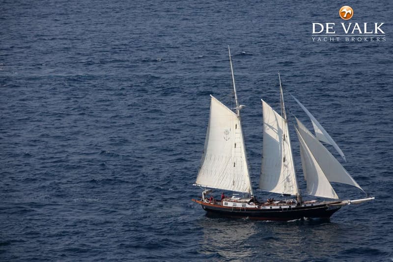 Een zeilschip met drie masten en witte zeilen vaart op een blauwe zee, met het logo van De Valk Yacht Brokers rechtsboven.