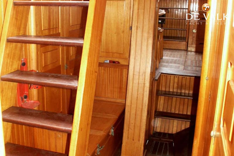 Een houten interieur met een trap, kasten en een gangpad, met een "DE VALK YACHT BROKER" watermerk.
