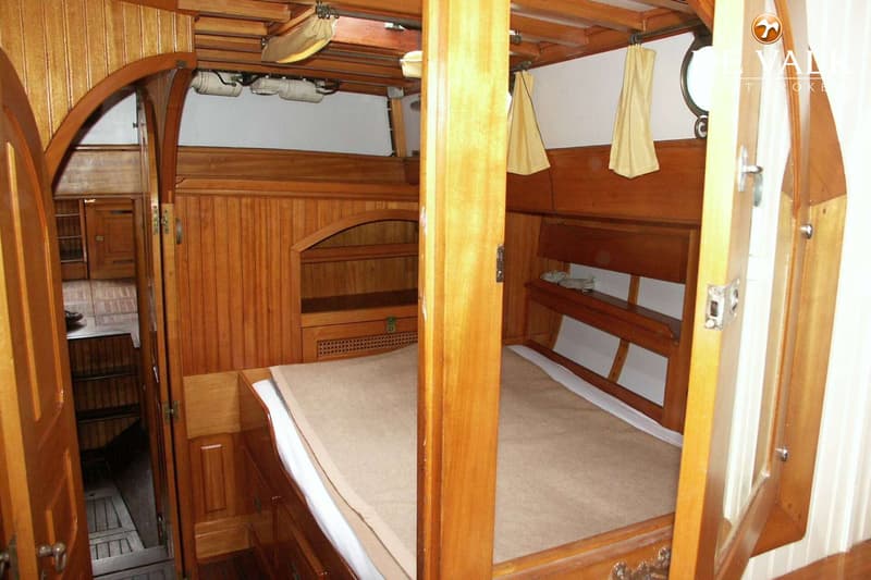 De foto toont een houten kajuitinterieur met een bed, opbergruimte en een doorgang naar een ander deel van de boot.