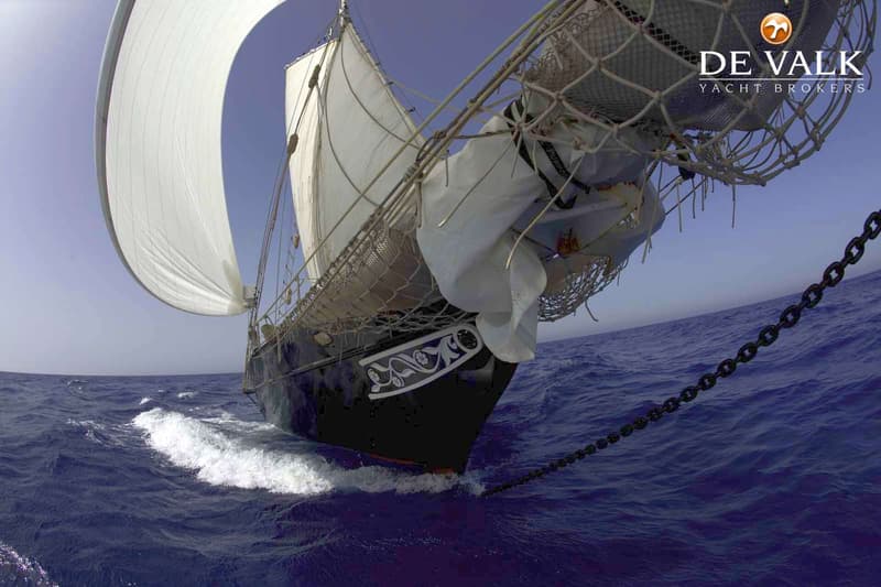 Een zeilschip met zwarte romp en witte zeilen vaart op een blauwe zee onder een heldere hemel.