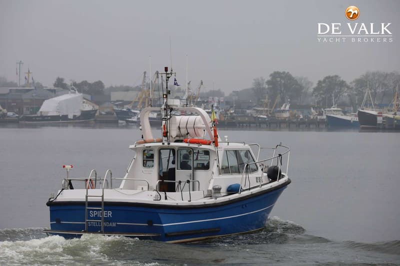Een blauw-witte boot met de naam "SPIDER" en "STELLENDAM" vaart op water, met een haven op de achtergrond.