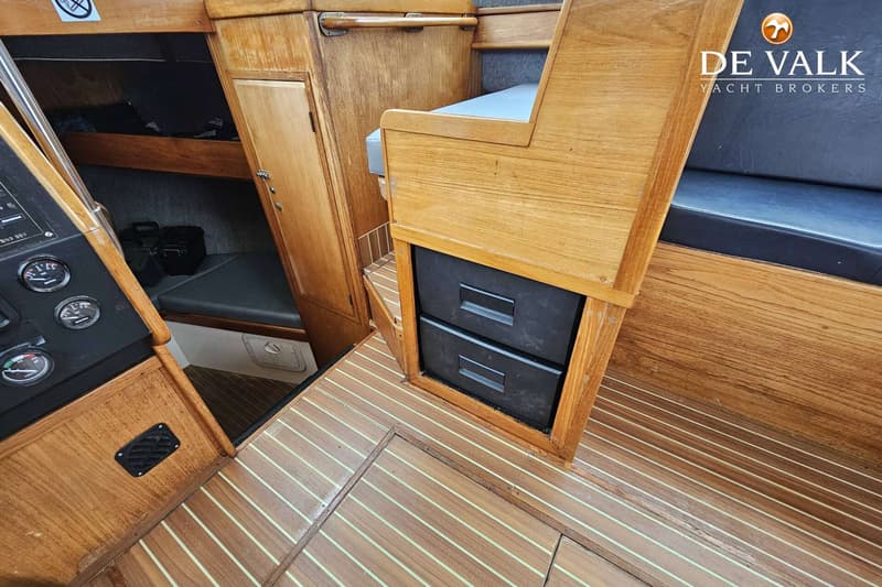 Een interieur van een boot met houten panelen, een donkere vloer met lichte strepen, en een zitbank met twee zwarte lades…