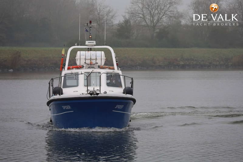 Een blauw-witte motorboot met de naam "SPIDER" vaart op een waterweg met een groene oever en kale bomen op de achtergrond.