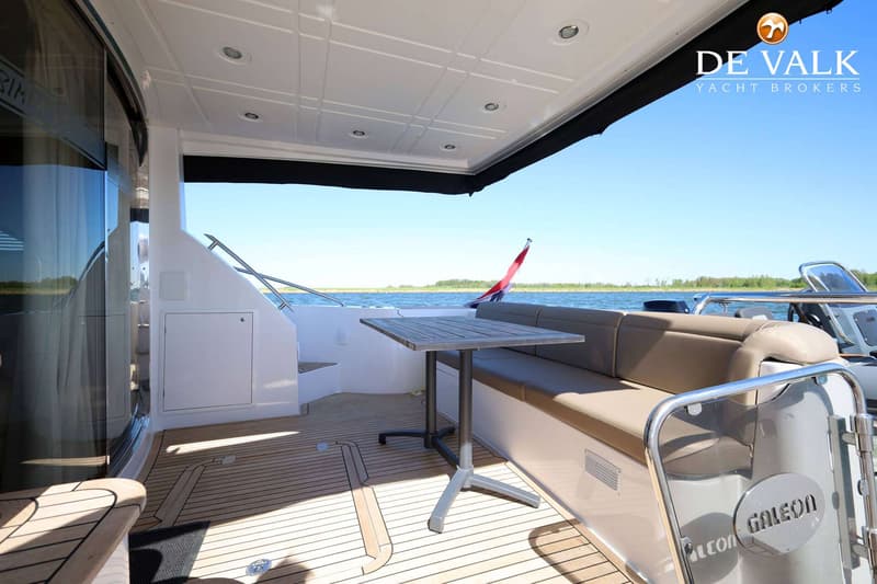 Een achterdek van een boot met een houten vloer, een bank met beige kussens, een tafel en uitzicht op water en land.