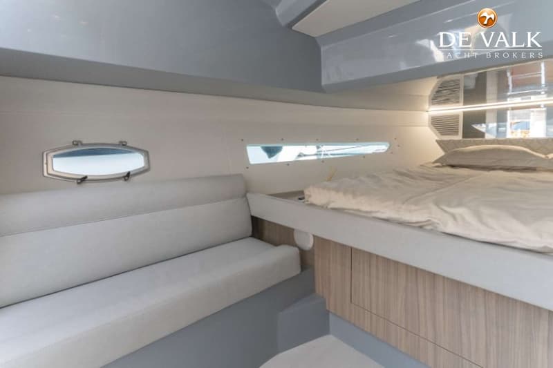 De foto toont een kajuit met een bed met wit beddengoed, een bank met grijze kussens, een patrijspoort en een langwerpig…