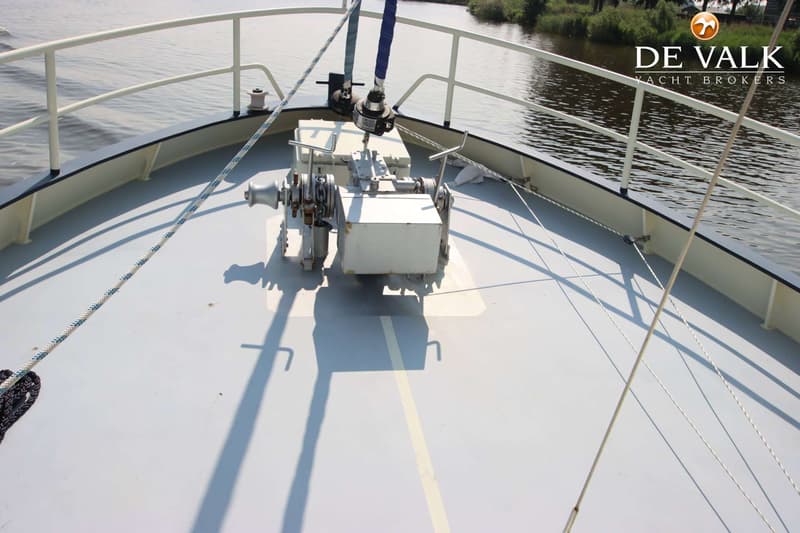 De foto toont het voordek van een boot met een ankerlier, reling, touwen en water op de achtergrond.