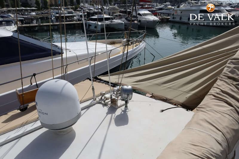 Een witte boot met een beige zeilhoes en een witte satellietkoepel in een haven vol met andere boten.