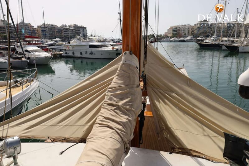 Een boot met opgerolde zeilen en een houten mast ligt in een haven vol met andere boten en gebouwen op de achtergrond.