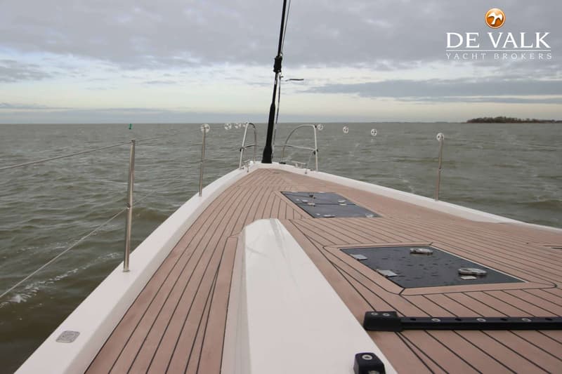 De foto toont het voordek van een zeilboot met een houten dek, reling en luiken, varend op een waterlichaam onder een…