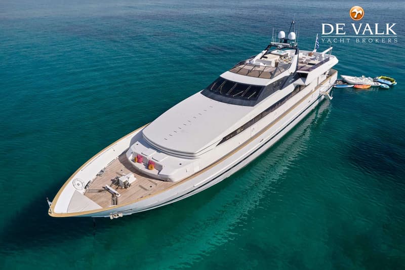 Een witte superjacht met een houten dek ligt in helder turquoise water, met een logo van "DE VALK YACHT BROKERS" rechtsboven.
