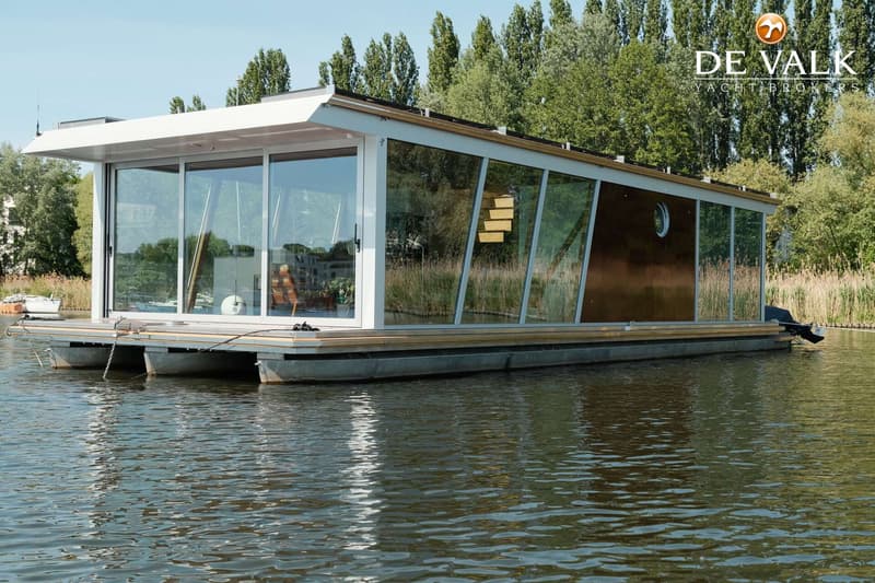 Een moderne woonboot met grote ramen en een plat dak ligt op het water, omringd door riet en bomen.