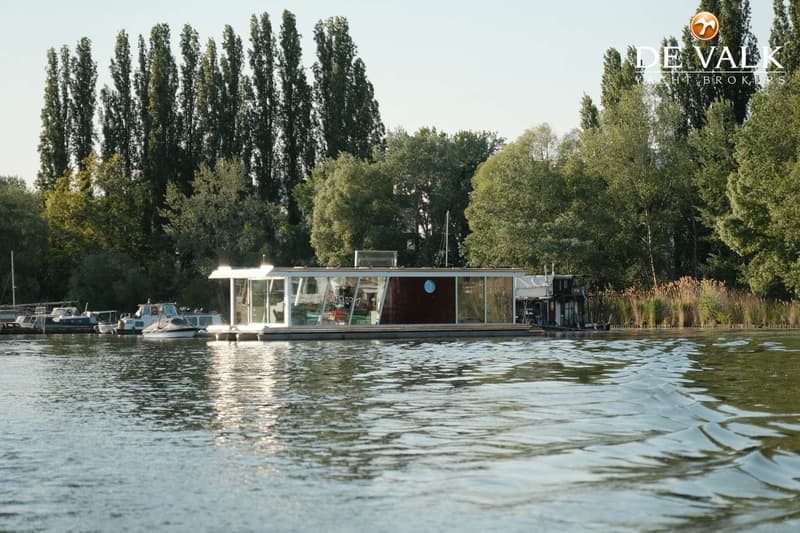 Een moderne woonboot met grote ramen en een bruine zijwand ligt op het water, met bomen en riet op de achtergrond.