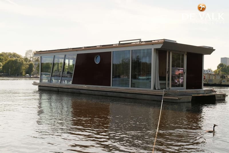 Een modern woonboot met grote ramen en een patrijspoort, drijvend op water met een gans in de voorgrond.