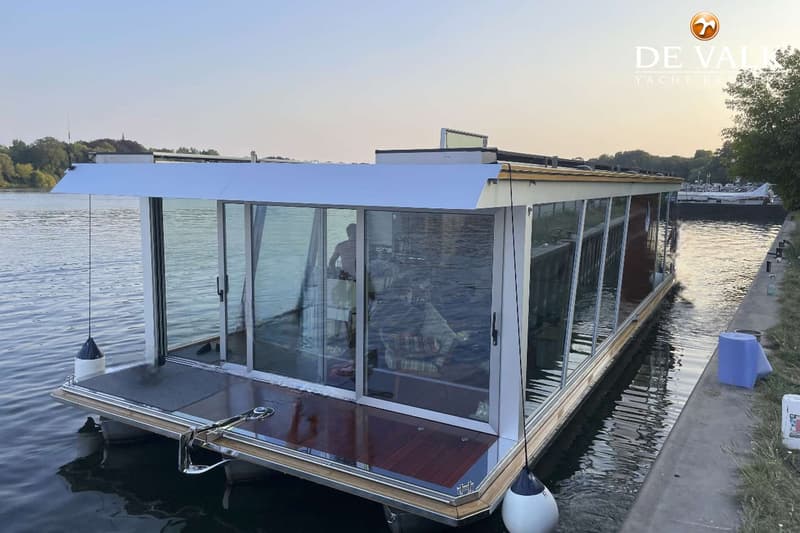Een moderne woonboot met grote glazen wanden ligt aangemeerd aan een kade, met een persoon zichtbaar binnenin.