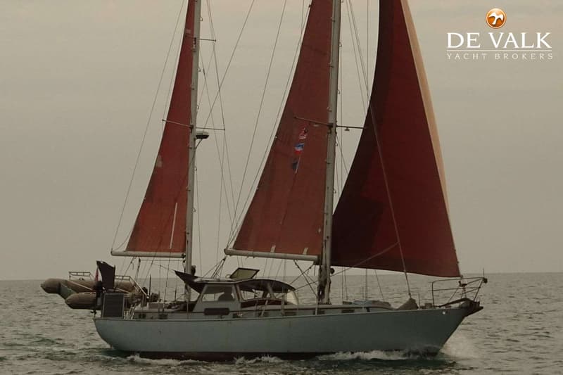 Een zeilboot met twee masten en rode zeilen vaart op zee, met een logo van "DE VALK YACHT BROKERS" rechtsboven.