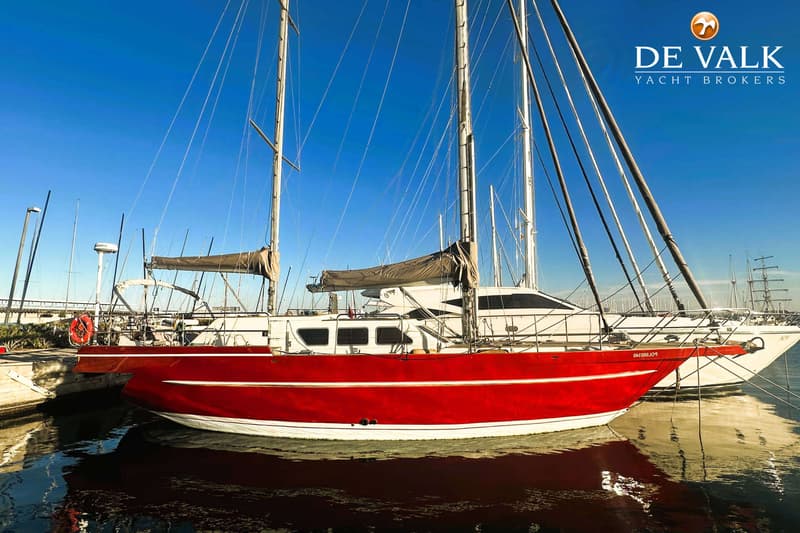 Een rode zeilboot met witte strepen ligt in het water, met een blauwe lucht erboven en andere boten op de achtergrond.