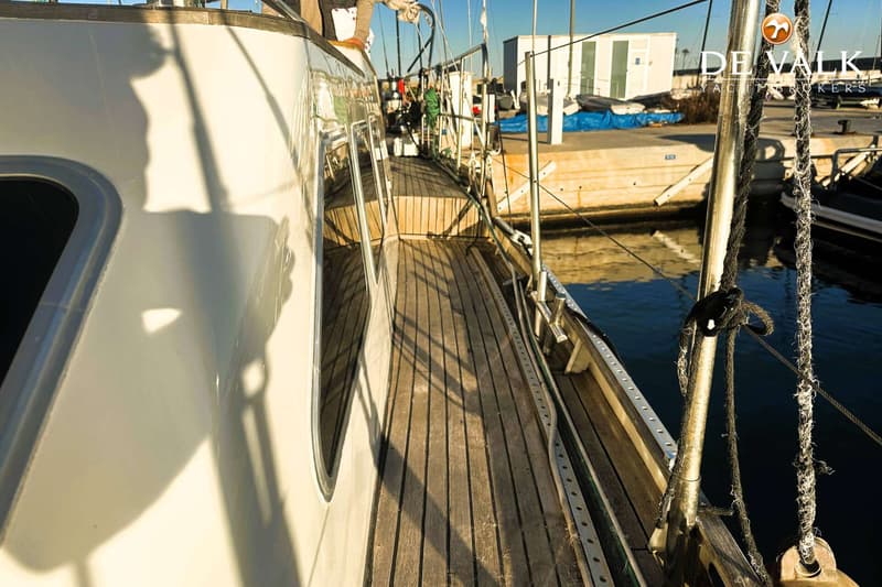 Een zijaanzicht van een boot met een houten dek, liggend aan een kade met water en gebouwen op de achtergrond.